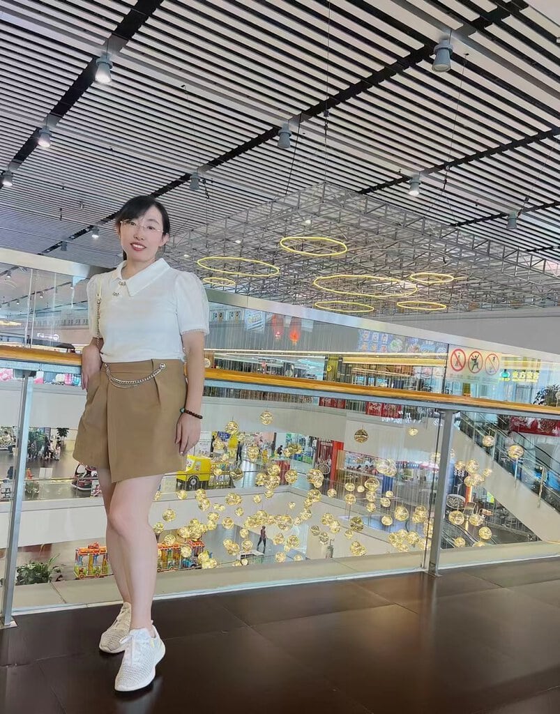 A person wearing a white top and brown skirt stands on a mall balcony. Behind them, decorative lights hang from the ceiling, and various stores are visible in the background.